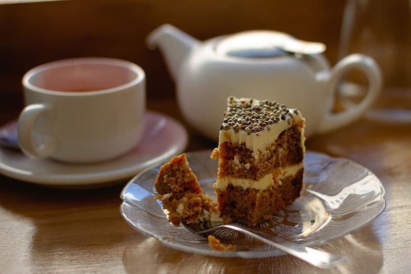 A slice of layered cake with frosting and sprinkles, alongside a teacup and teapot on a wooden table.
