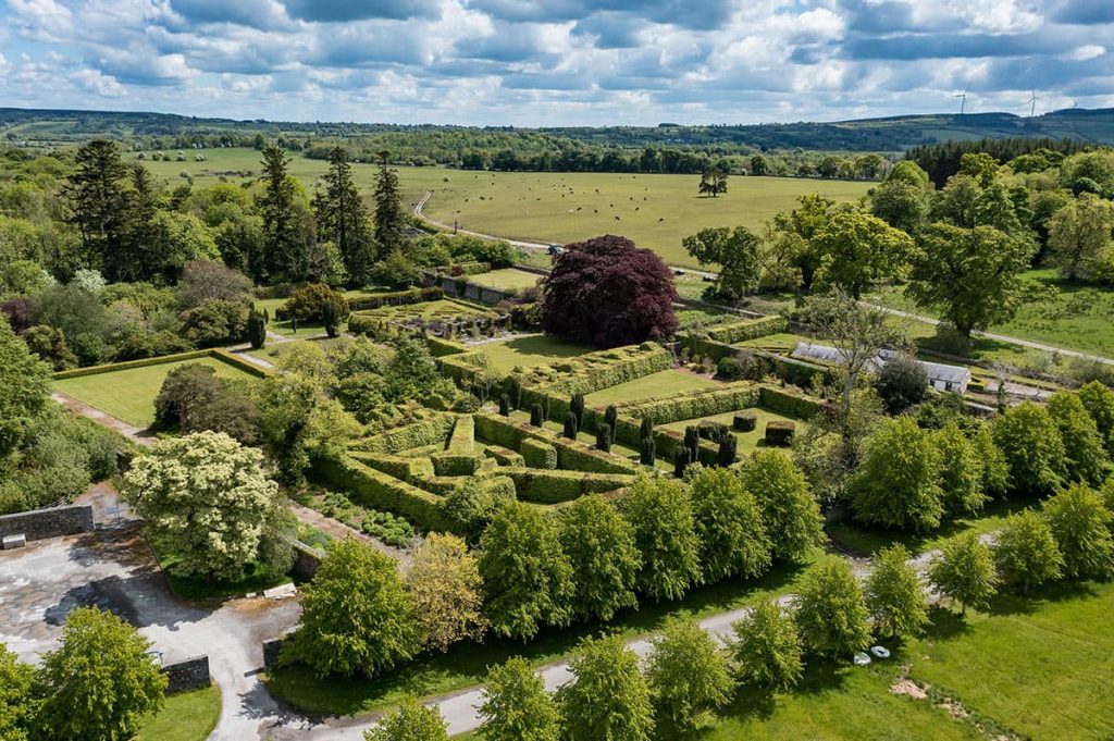 Aerial view of a formal garden with symmetrical hedges, a large central tree, and surrounding green fields under a cloudy sky.