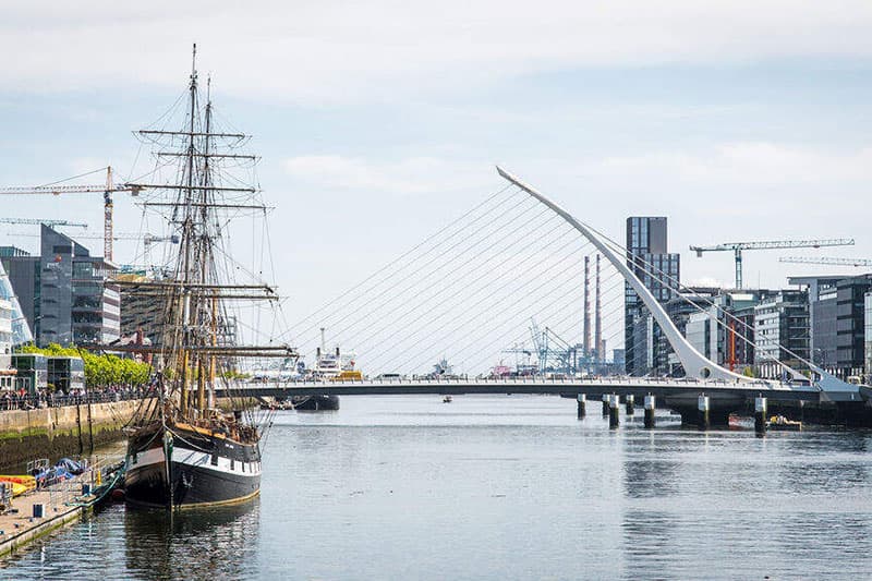 A tall ship docked along a river with modern buildings and cranes nearby, and a distinctive white cable-stayed bridge in the background.