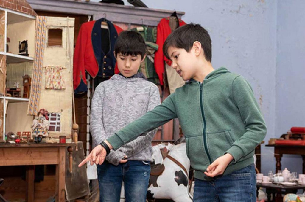 Two boys interact in a vintage-themed room filled with toys and historical clothing, showcasing their curiosity and engagement.