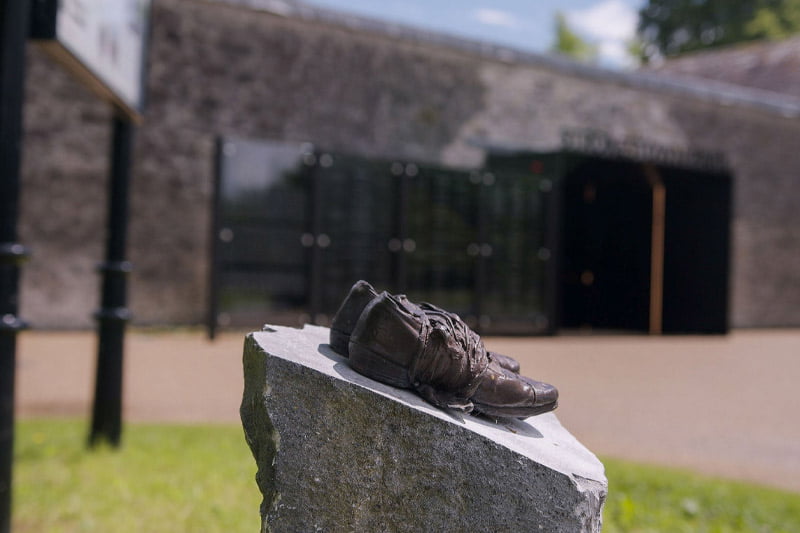 Bronze shoes sculpture on a stone pedestal with a blurred background of a historical building and entrance.