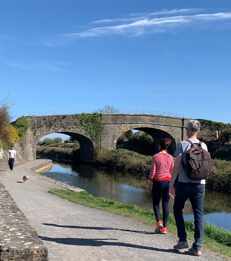 Two people stroll along a riverbank path under a historic stone bridge, with a sunny blue sky and a small dog walking nearby.