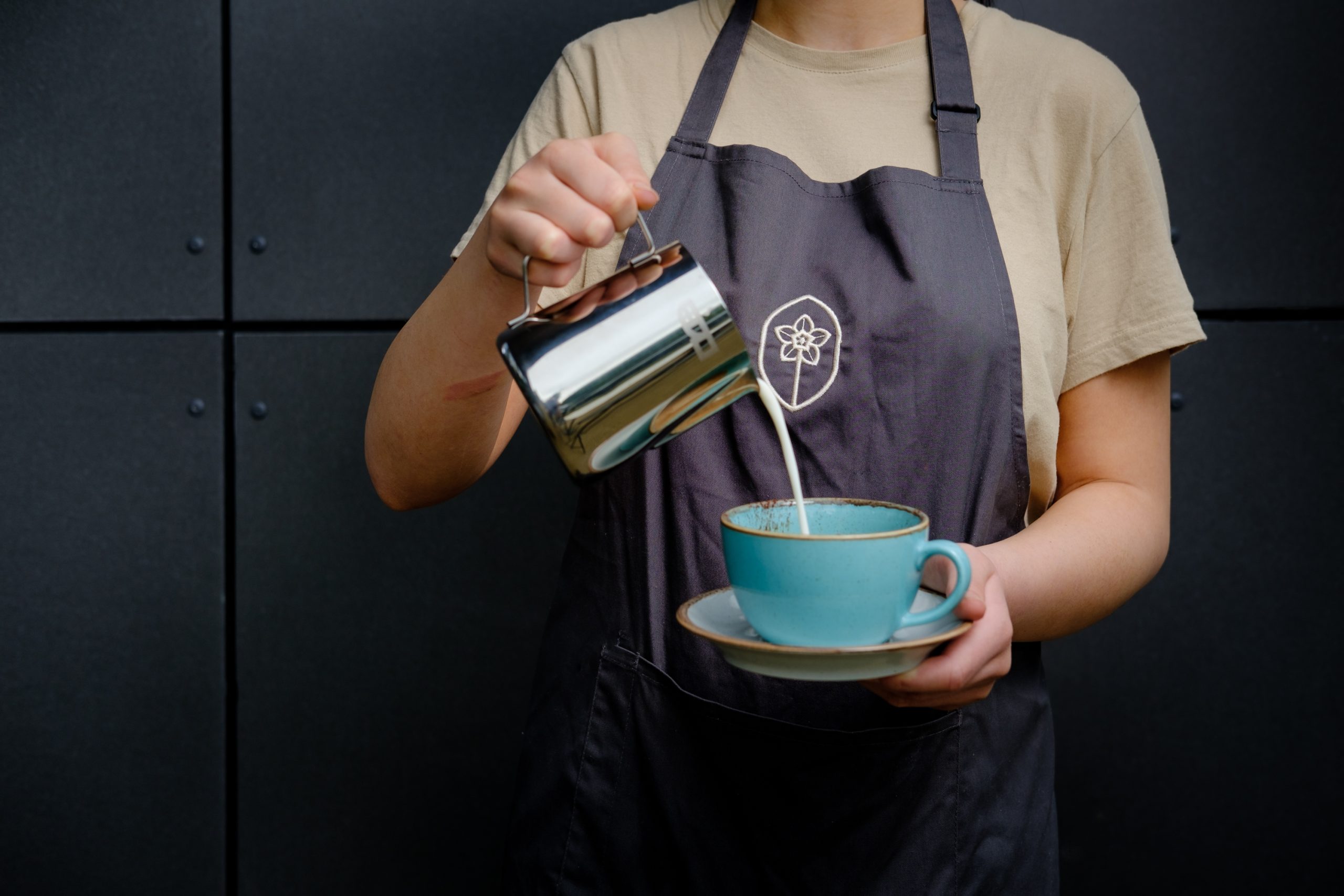Waiting Staff dressed in beige tshirt and graphite apron featuring an embroidered illustration of a potato flower, pouring milk from a steel jug into a teal coffee cup.