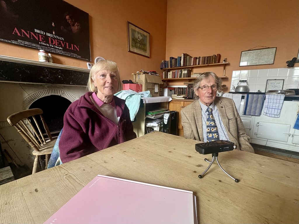 Irish Heritage Trust Volunteer and Mr. Anthony Owens sitting at wooden table containing recording equipment in Catherine's Kitchen, Strokestown Park House.